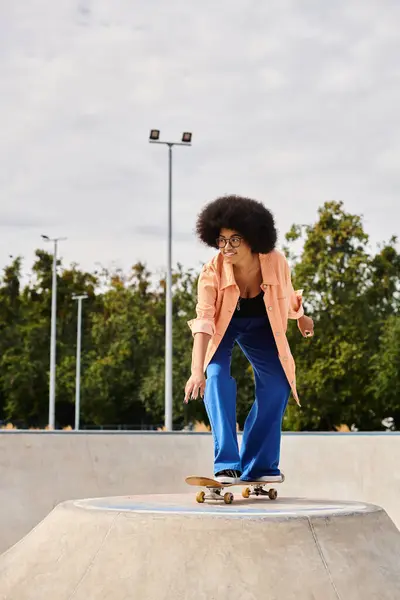 A young African American woman with curly hair rides a skateboard on top of a cement ramp in an urban skate park. — Stock Photo