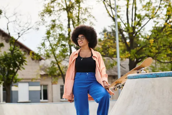 Jeune femme afro-américaine aux cheveux bouclés se tient à côté de skateboard sur une rampe de skateboard dans un parc de skate extérieur dynamique. — Photo de stock