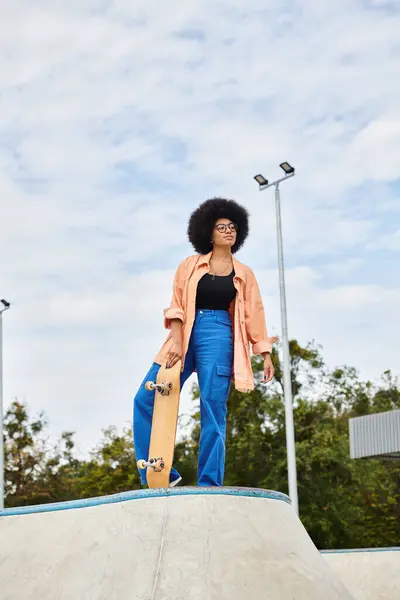 Eine junge Afroamerikanerin hält stolz ein Skateboard an der Spitze einer Rampe in einem Skatepark im Freien. — Stockfoto