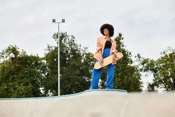A young African American woman with curly hair confidently stands atop a skateboard ramp at an outdoor skate park. — Stock Photo