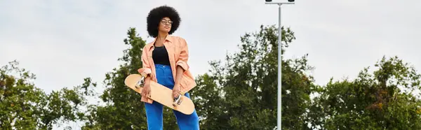 Молодая афроамериканка с вьющимися волосами изящно стоит на скейтборде в ярком парке. — стоковое фото