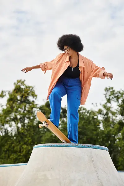 A young African American woman with curly hair rides a skateboard on top of a cement ramp in an outdoor skate park. — Stock Photo