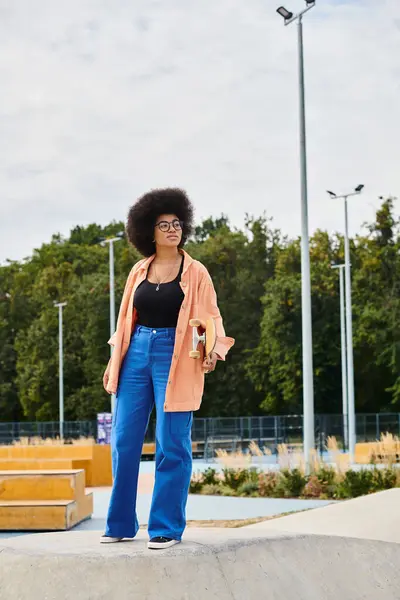 Une jeune Afro-Américaine aux cheveux bouclés se tient en confiance sur une rampe de skateboard dans un skate park, prête pour son prochain mouvement. — Photo de stock
