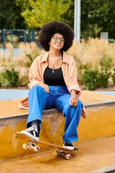 Una joven afroamericana con el pelo rizado se sienta en una repisa sosteniendo un monopatín en el parque de skate. - foto de stock