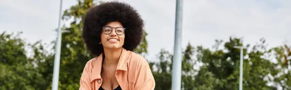 A stylish young African American woman with curly hair wearing glasses and a pink shirt skateboarding outdoors in a skate park. — Stock Photo