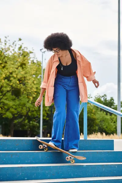 Una talentosa joven afroamericana con el pelo rizado monta hábilmente su monopatín por un tramo de escaleras en un parque de skate. - foto de stock