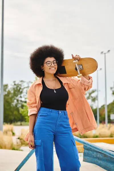 Une jeune Afro-Américaine avec un afro tient en toute confiance une planche à roulettes dans un parc de skate dynamique. — Photo de stock
