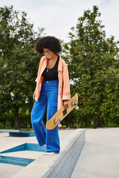 A young African American woman with curly hair stands confidently on a ledge with her skateboard in a skate park. — Stock Photo