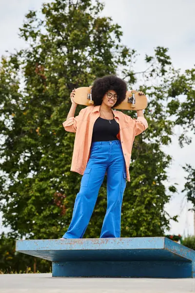 African American woman with curly hair strikes a pose holding a skateboard on a blue platform in a skate park. — Stock Photo