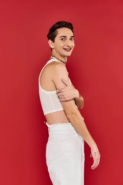 Joyeux bel homme gay avec des accessoires élégants en tenue blanche en regardant la caméra sur fond rouge — Photo de stock