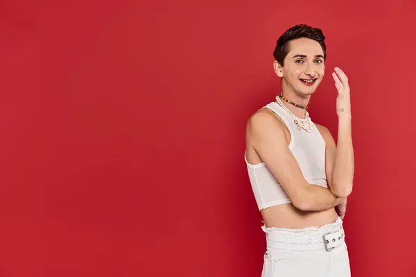 Joyeux bel homme gay avec des accessoires élégants en tenue blanche en regardant la caméra sur fond rouge — Photo de stock