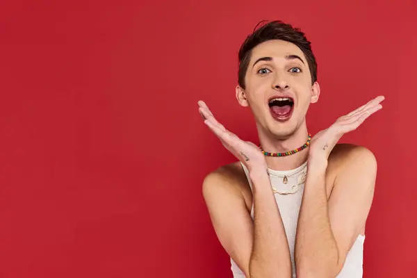 Cheerful handsome gay man with stylish accessories in white attire looking at camera on red backdrop — Stock Photo