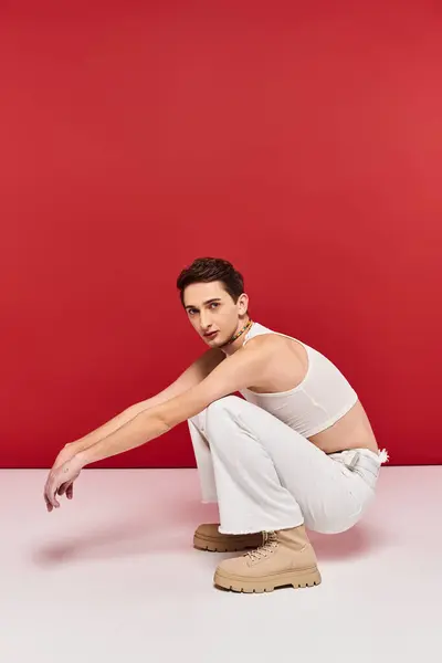Atractivo joven gay hombre con elegante accesorios en blanco atuendo mirando la cámara en rojo telón de fondo - foto de stock