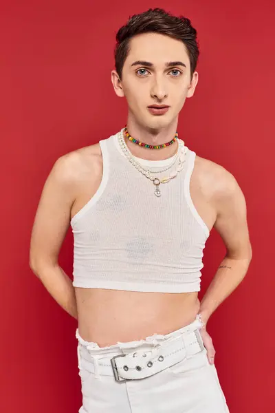 Atractivo joven gay hombre con elegante accesorios en blanco atuendo mirando la cámara en rojo telón de fondo - foto de stock