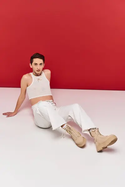Beau androgyne homme en tenue décontractée assis sur le sol et regardant la caméra sur fond rouge — Photo de stock