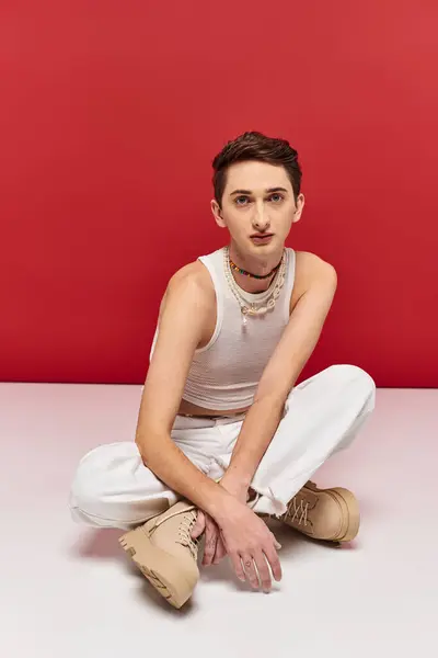Beau androgyne homme en tenue décontractée assis sur le sol et regardant la caméra sur fond rouge — Photo de stock