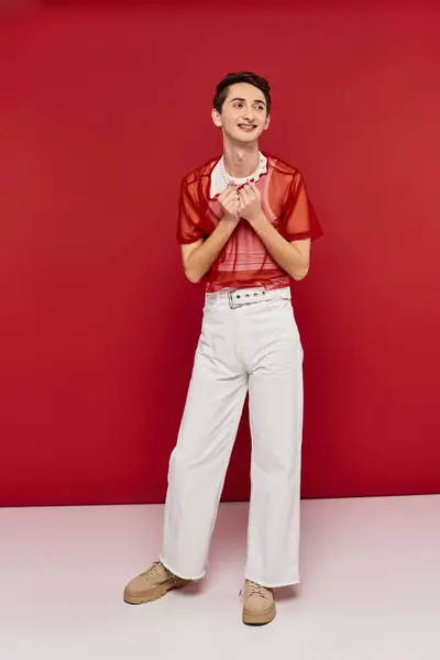 Cheerful androgynous man in white attire with red fishnet posing on red backdrop and looking away — Stock Photo