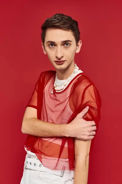 Attractive voguish androgynous man with stylish red fishnet looking at camera on red backdrop — Stock Photo