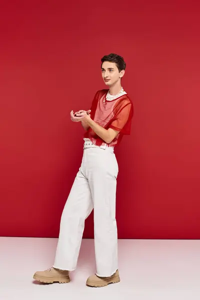Красивий андрогінний чоловік у білому одязі з червоною сіткою, що позує на червоному тлі і дивиться в сторону — стокове фото