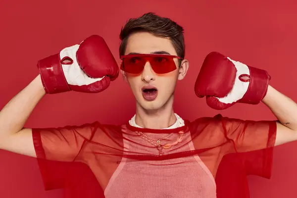 Fashionable androgynous man with red fishnet posing actively with boxing gloves and looking away — Stock Photo