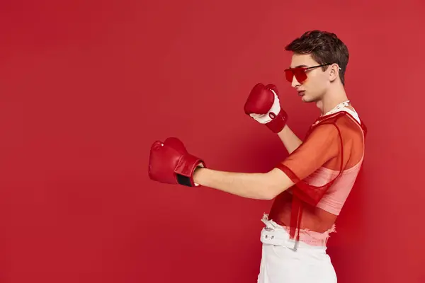 Hombre andrógino de moda con red roja posando activamente con guantes de boxeo y mirando hacia otro lado - foto de stock