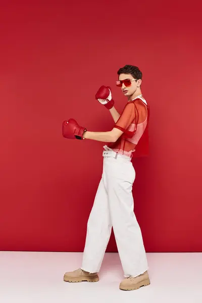 Bel homme gay avec des lunettes de soleil et résille rouge posant avec des gants de boxe et regardant la caméra — Photo de stock