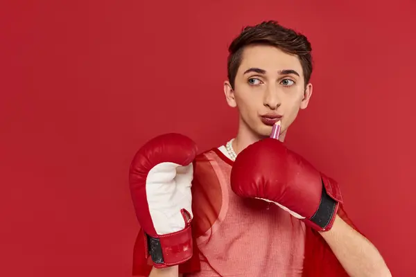 Handsome stylish androgynous man with boxing gloves posing with lipstick in hands and looking away — Stock Photo