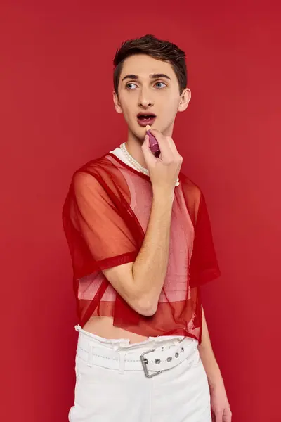 Sophisticated androgynous man with red fishnet holding lipstick and looking away on red backdrop — Stock Photo