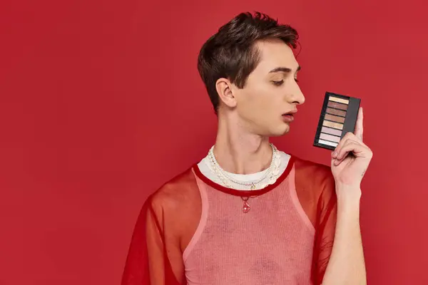 Hombre andrógino de moda con elegante red de pescados rojo sosteniendo la paleta de sombras de los ojos y mirando hacia otro lado - foto de stock