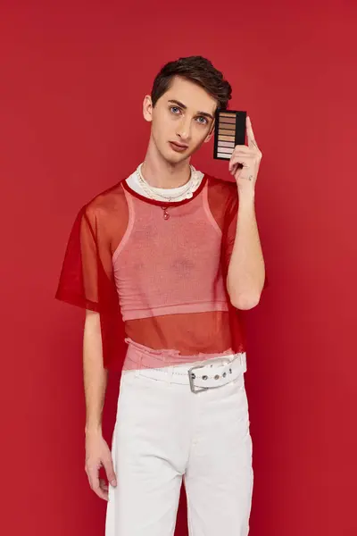 Handsome androgynous man with red fishnet holding eye shadows palette and looking at camera — Stock Photo