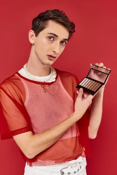 Bel homme androgyne avec résille rouge tenant la palette des ombres à paupières et regardant la caméra — Photo de stock