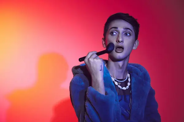 Fashionable androgynous man in stylish attire using makeup brush and posing on vibrant backdrop — Stock Photo