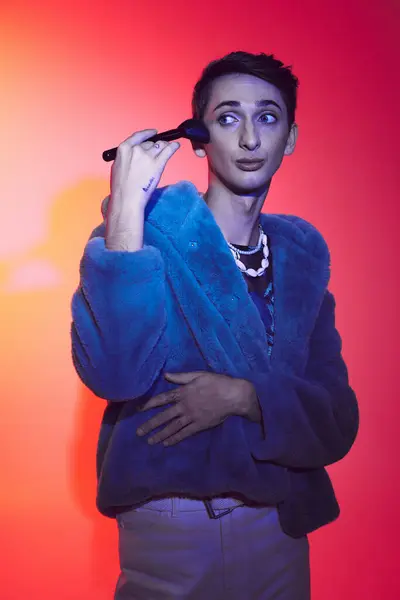 Hombre andrógino sofisticado en traje elegante usando cepillo de maquillaje y posando sobre un vibrante telón de fondo - foto de stock