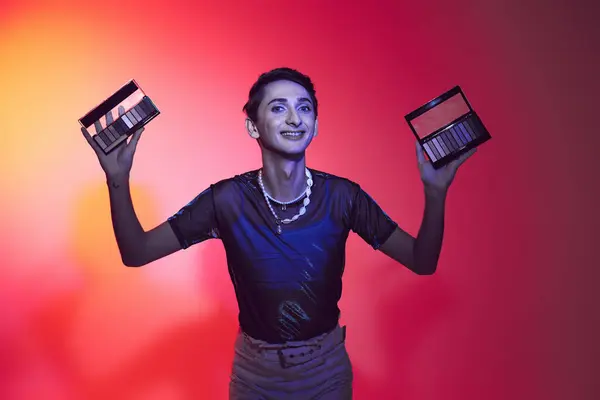 Tempting gay man in casual attire using eyeshadows palette and looking at camera on vibrant backdrop — Stock Photo