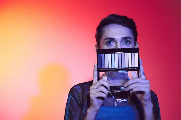 Enticing gay man in casual attire using eyeshadows palette and looking at camera on vibrant backdrop — Stock Photo