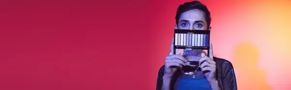 Chic gay man in cozy attire using eyeshadows palette and looking at camera on vivid backdrop, banner — Stock Photo