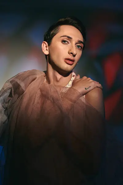 Tentador andrógino fashionista com cabelo escuro em traje pastel olhando para a câmera em pano de fundo vívido — Fotografia de Stock