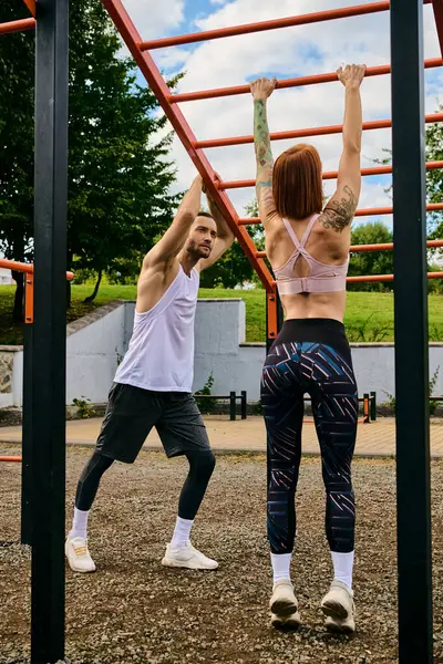 A determined woman, in sportswear, execute pull-ups side by side with a personal trainer outdoors. — Stock Photo