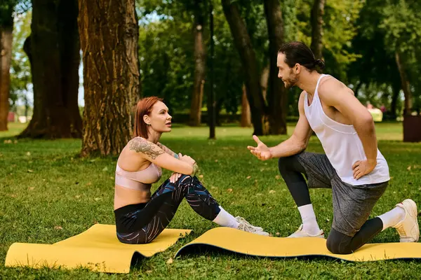 Un hombre y una mujer en ropa deportiva hacen ejercicio sobre colchonetas de yoga en un parque, guiados por un entrenador personal. - foto de stock