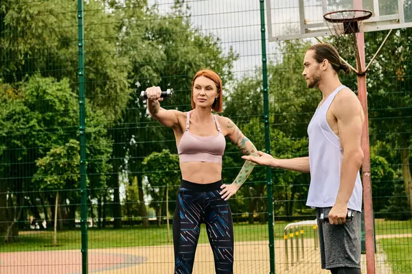 A determined woman in sportswear exercising with dumbbell near personal trainer outdoors — Stock Photo