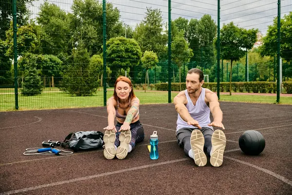 Ein entschlossener Mann und eine entschlossene Frau, beide in Sportbekleidung, sitzen zusammen auf einem Basketballplatz und erreichen ihre Fitness-Ziele. — Stockfoto