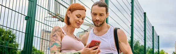 Un hombre y una mujer en ropa deportiva se centran en una pantalla de teléfono celular, posiblemente siguiendo un plan de entrenamiento guiado por un entrenador personal. - foto de stock