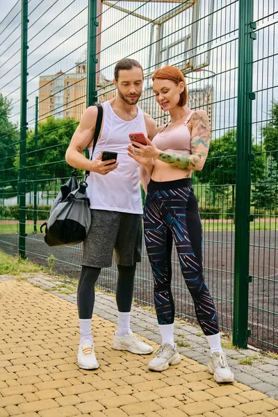 Un hombre y una mujer en ropa deportiva se paran lado a lado al aire libre, sosteniendo teléfonos inteligentes - foto de stock