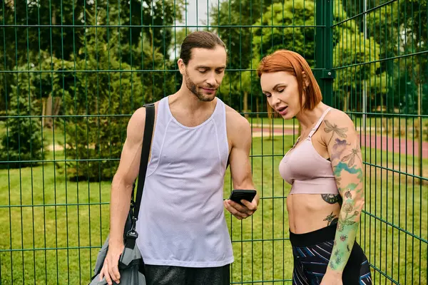 Um homem e uma mulher determinados no sportswear estão na frente de uma cerca, usando o smartphone — Fotografia de Stock