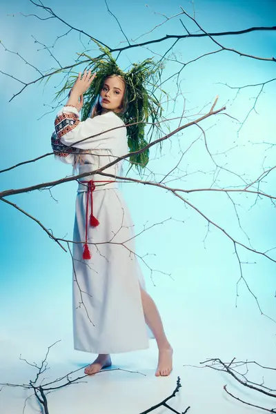 Une jeune femme en robe blanche et tenue traditionnelle se tient gracieusement devant un arbre mystique dans un décor de studio. — Photo de stock