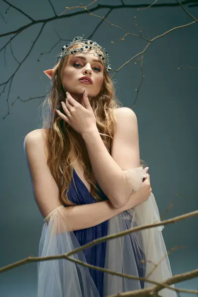 Uma jovem mulher, assemelhando-se a uma princesa duende conto de fadas, fica graciosamente em um vestido azul na frente de uma árvore majestosa. — Fotografia de Stock