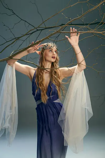 Uma jovem mulher em um vestido azul fica graciosamente, segurando um galho de árvore em um estúdio. Ela transpira uma essência de conto de fadas, semelhante a uma princesa duende. — Fotografia de Stock