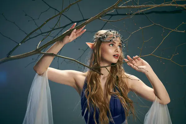 Una joven vestida con un vestido azul fluido se levanta con gracia junto a un majestuoso árbol en un entorno de fantasía. - foto de stock