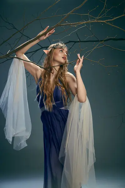 A young woman in a blue dress resembling an elf princess, delicately holds a branch in a studio setting. — Stock Photo