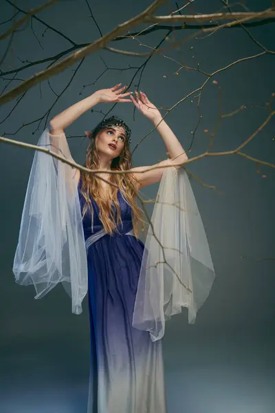 Una joven con un vestido azul y blanco se levanta con gracia junto a un árbol en un entorno de hadas y fantasía inspirada. - foto de stock